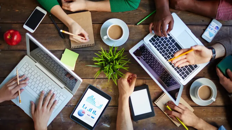 Teamwork around a table with laptops, tablets, and coffee cups.